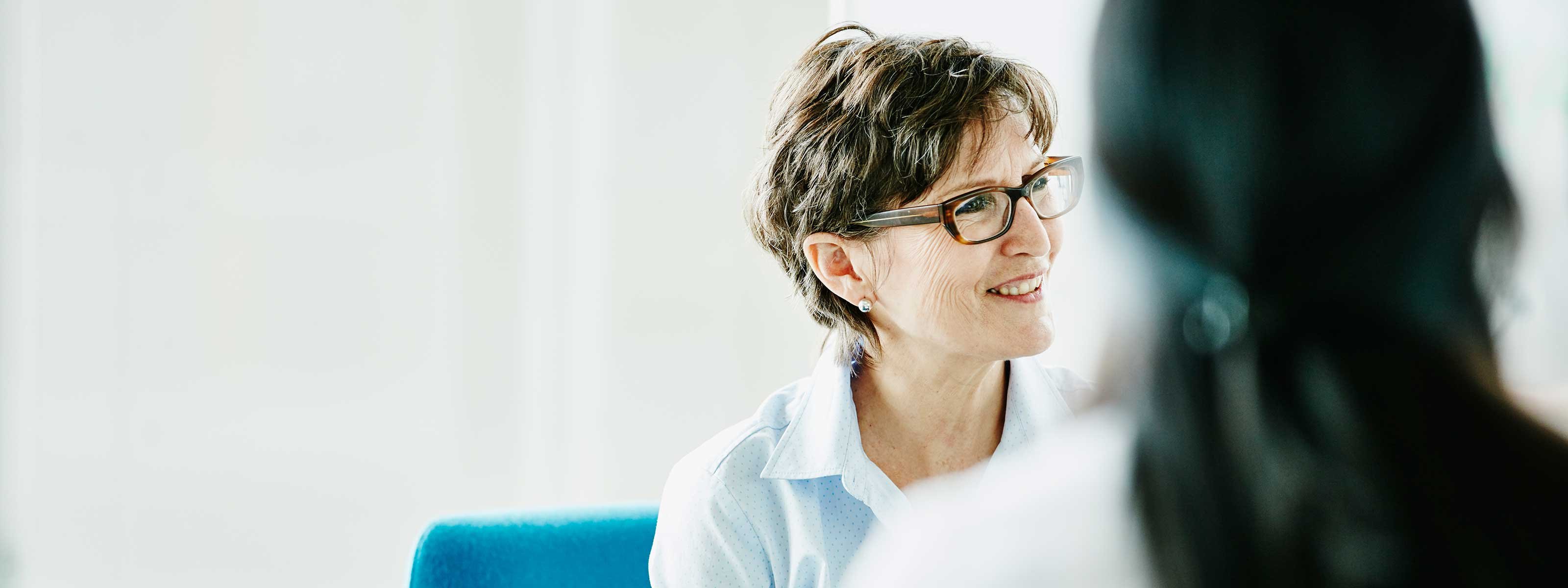 Person wearing glasses in a white coat speaking with another person in a bright indoor setting